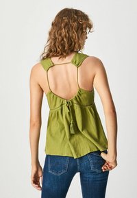 Top verde senza maniche con tessuto testurizzato, schiena scoperta, legatura regolabile con nappina e spalline larghe. Abbinato a jeans in denim.