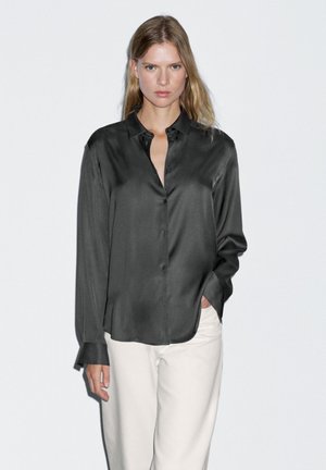 Blusa in satin grigio scuro con maniche lunghe e chiusura a bottoni, abbinata a pantaloni di colore chiaro. Texture liscia e vestibilità rilassata.
