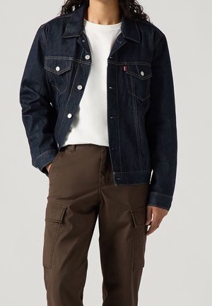 Personne portant une veste en jean bleu foncé sur une chemise blanche avec un pantalon cargo marron, debout avec une main dans la poche.