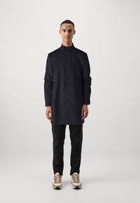 Cappotto in misto lana blu navy con colletto alto, chiusura a bottoni e tasche a patta; abbinato a jeans scuri e sneakers beige.