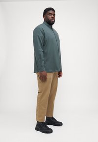 Camisa verde de botones, pantalones marrones y botas negras resistentes. Incluye mangas largas, un bolsillo en el pecho y un corte relajado.