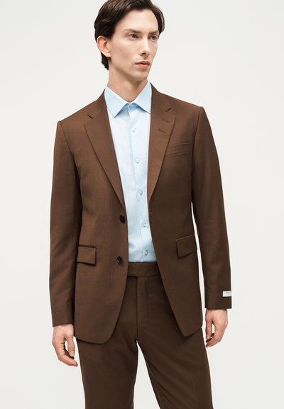 Άνδρας που φοράει καφέ κοστούμι ραμμένο στα μέτρα με μαύρα κουμπιά και γαλάζιο πουκάμισο, στέκεται μπροστά σε λευκό φόντο.