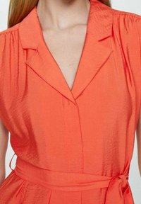 Koralowa sukienka z kołnierzykiem, krótkimi bufkami i paskiem w talii; gładka, lekko fakturowana tkanina.