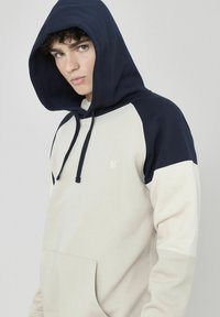 Sweat à capuche avec un corps beige et des manches raglan bleu marine. Fabriqué en tissu doux, avec une poche avant et un capuchon à cordon.