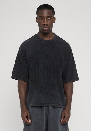 Jeune homme noir portant un t-shirt oversize noir délavé et ample ainsi qu'un short assorti, debout devant un fond clair uni.