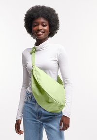 Borsa a tracolla verde in tessuto con una superficie liscia, forma curva, tracolla regolabile e branding discreto, indossata a tracolla.
