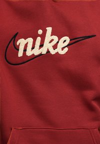 Punainen paita, jossa suuri, kirjailtu kermanvärinen "nike"-logo ja musta swoosh-logo sen alla. Pehmeä kangas ja etutasku näkyvissä.