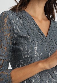 Top en dentelle grise avec un décolleté en V et des manches trois quarts, présentant des motifs floraux et une surface texturée.