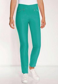 Türkise, enganliegende Hose mit glatter Textur, die zwei mit Reisverschlüssen ausgestattete Vordertaschen und einen hohen Bund hat. Kombiniert mit weißen Turnschuhen.