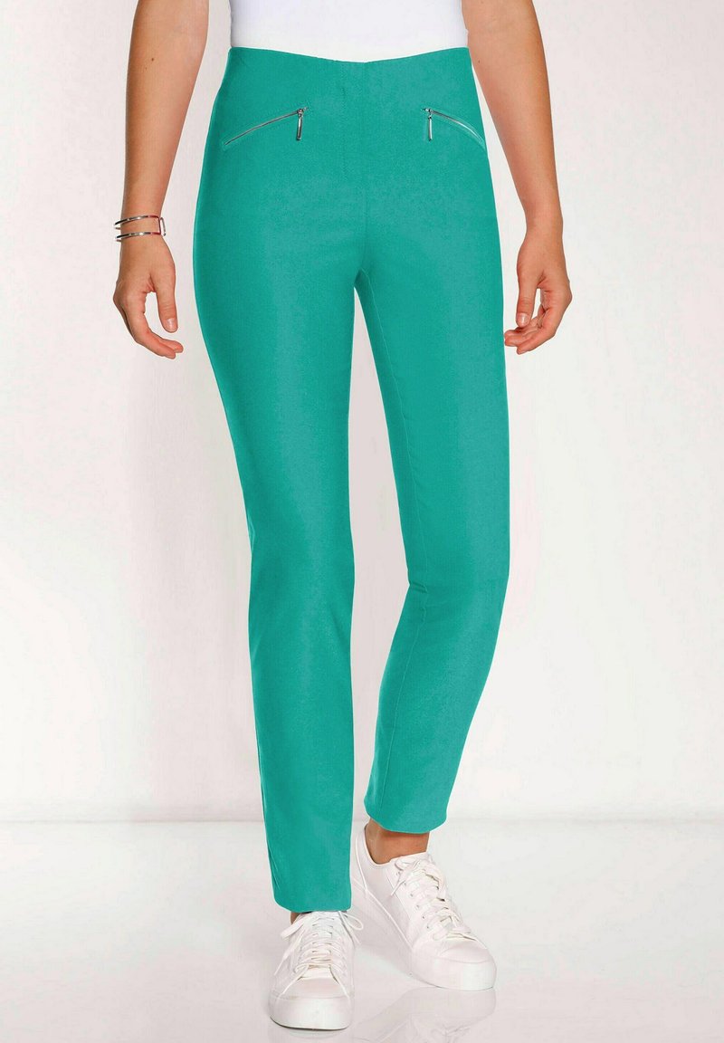 Türkise, enganliegende Hose mit glatter Textur, die zwei mit Reisverschlüssen ausgestattete Vordertaschen und einen hohen Bund hat. Kombiniert mit weißen Turnschuhen.