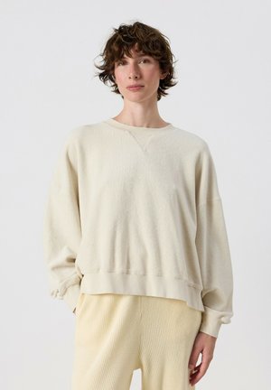 Persona con capelli corti e ricci castani indossa un maglione largo color avorio e pantaloni beige chiaro texturizzati, in piedi davanti a uno sfondo semplice.