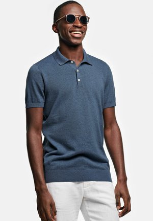 Lachende man die een donkerblauw poloshirt met korte mouwen, witte broek en bruine zonnebril draagt tegen een effen witte achtergrond.