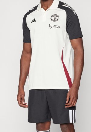 Mężczyzna w białej i czarnej koszulce polo Manchester United z czerwonymi bocznymi paskami, czarnych szortach Adidas z białymi paskami i skarpetkach.