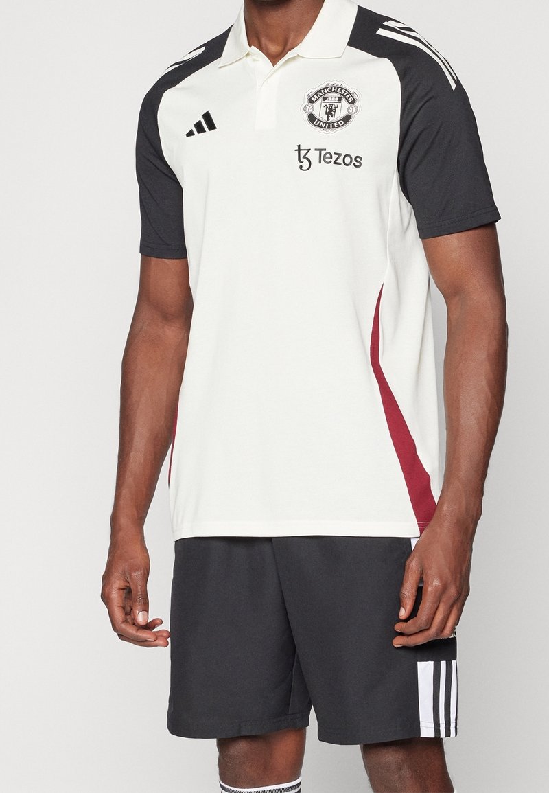 Homme portant un polo Manchester United blanc et noir avec des bandes rouges sur le côté, un short Adidas noir avec des bandes blanches et des chaussettes.