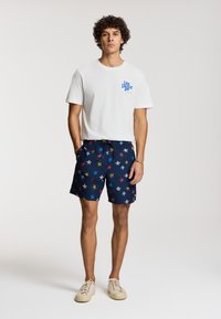Fehér pamut póló kék felirattal, tengerészkék fürdőshorts-ban, amely színes csillaghal mintázattal rendelkezik. Lazacipő krémszínben.