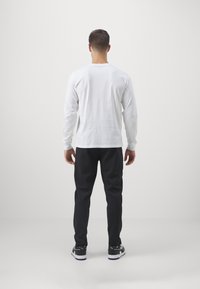 Man staan met zijn rug naar de camera, gekleed in een wit lange mouwen shirt, zwarte broeken en zwarte Puma sneakers tegen een effen lichte achtergrond.