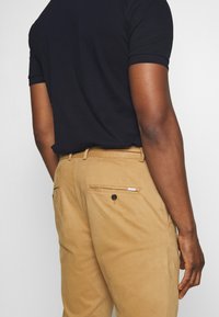 Pantalons khaki avec une coupe droite, deux poches arrière avec des boutons, et une texture de tissu lisse ; associés à un T-shirt noir.