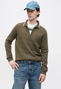 Jovem vestindo um boné azul, suéter verde oliva com meia abertura sobre uma camisa listrada e jeans azuis, em pé contra um fundo branco liso.