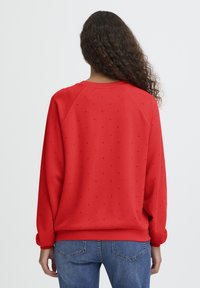 Een vrouw met lang, krullend haar, gekleed in een rode sweatshirt met kleine versieringen en een blauwe spijkerbroek, gezien van achteren tegen een effen achtergrond.