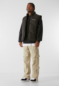 Opgevulde zwarte vest met hoge kraag, gecombineerd met baggy lichtbeige cargobroeken met meerdere zakken. Witte en zwarte sneakers maken de look compleet.