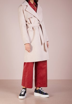 Personne portant un manteau beige ceinturé sur un pantalon large en velours côtelé rouge et des baskets basses noir et blanc, debout devant un fond uni.