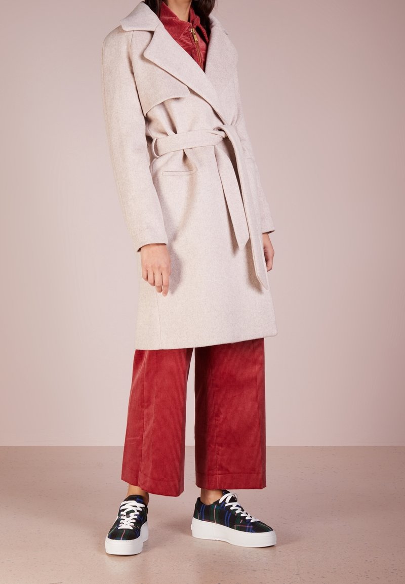 Personne portant un manteau beige ceinturé sur un pantalon large en velours côtelé rouge et des baskets basses noir et blanc, debout devant un fond uni.