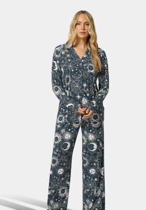 Femme aux longs cheveux portant un pyjama foncé avec des motifs blancs de soleil, lune et étoiles, debout les mains jointes devant sur un fond uni.