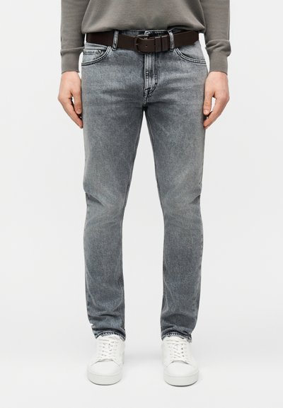 Άνδρας που φοράει ανοιχτό γκρι τζιν slim-fit, λευκά αθλητικά παπούτσια, γκρι μακρυμάνικο πουκάμισο και καφέ δερμάτινη ζώνη, στέκεται μπροστά σε λευκό φόντο.