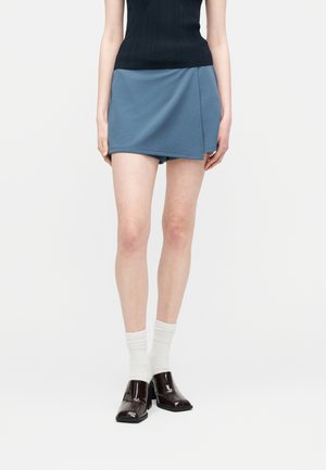 Kobieta ubrana w niebieską mini spódniczkę owijaną, czarny bezrękawnik, białe skarpetki i ciemnobrązowe błyszczące mokasyny, stojąca na jednolitym tle.