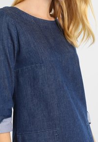 Top de mezclilla en azul oscuro con cuello redondo, mangas tres cuartos con pu�ños a rayas y un bolsillo frontal. Textura lisa.