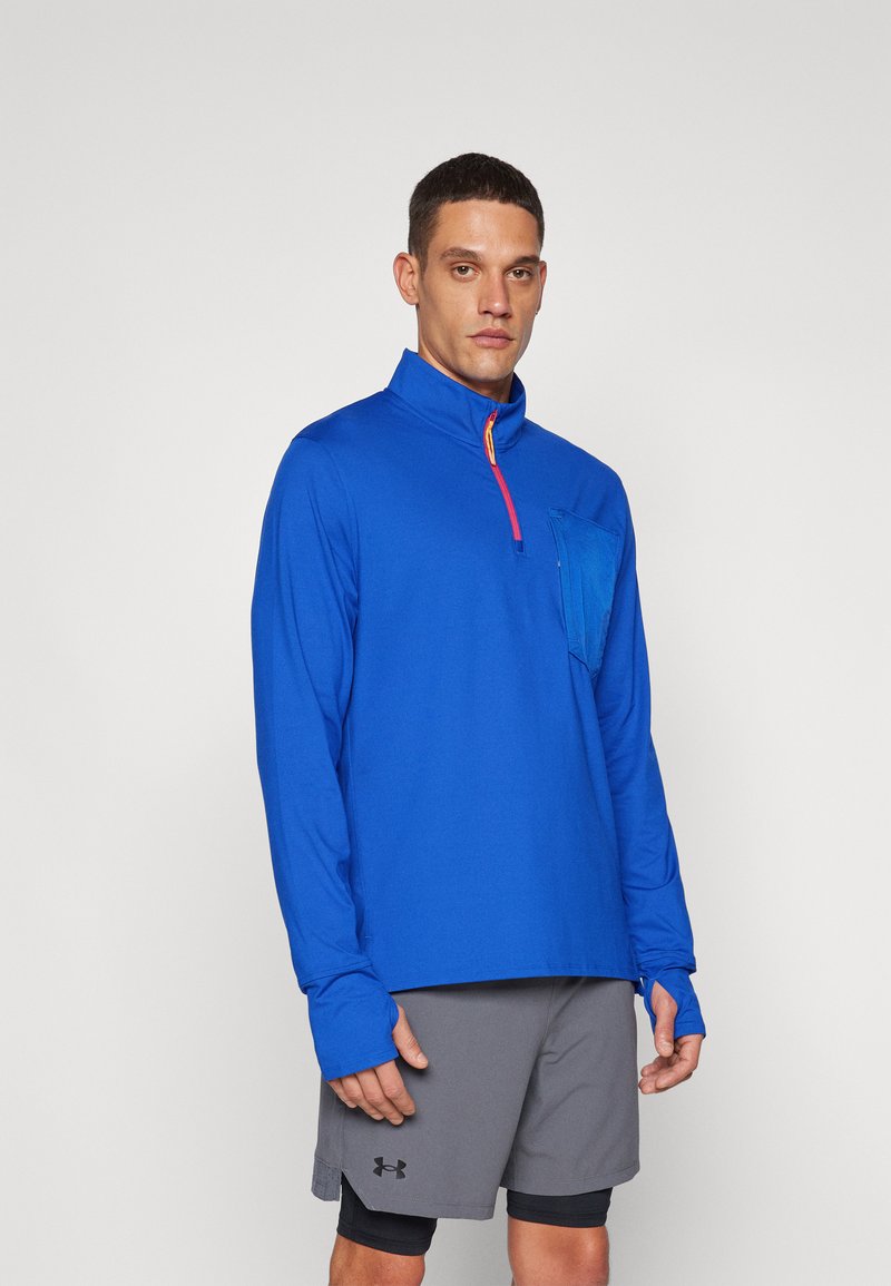 Blauer Langarmpullover mit Reißverschlusskragen, ausgestattet mit einer Fronttasche und Daumenlöchern. Kombiniert mit grauen Shorts. Glattes, leichtes Material.