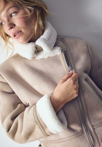 Žena v béžovej koženej bunde so strihanou srsťou a bielym fleece golierom a manžetami, drží zips blízko hrude a pozerá sa doľava.