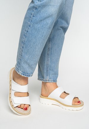 White slide sandals with dual wide straps, textured material, a cork footbed, and a ridged white sole. Pedicure features red polish.