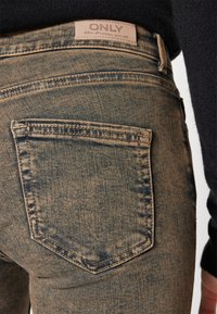 Jeans de mezclilla azul deslavado con un bolsillo trasero con detalles de costura y una etiqueta de la marca situada por encima de la línea de la cintura. Textura suave.