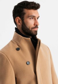 Abrigo de lana color camel con un collar alto, que presenta dos botones negros y una textura suave. Expuesto en un modelo sobre un fondo blanco.
