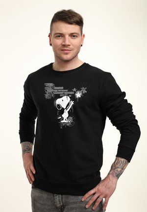 Hombre con tatuajes que lleva una sudadera negra con un dibujo de Snoopy alcanzando una estrella luminosa.