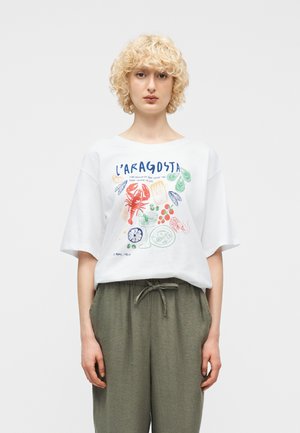 Άτομο με σγουρά ξανθά μαλλιά που φοράει λευκό ανοιχτό t-shirt με πολύχρωμες εικόνες θαλασσινών και ποτού και χακί παντελόνι με κορδόνι στη μέση.