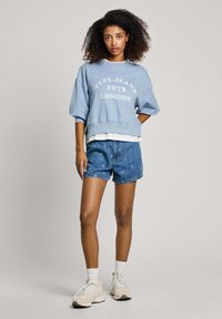 Blauwe sweatshirt met de opdruk "Pepe Jeans 1973 London", gecombineerd met denim short met bloemmotieven, aangevuld met witte sokken en sneakers.