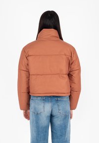 Brązowa kurtka puchowa z wysokim kołnierzem i subtelnym wzorem, o luźnym kroju z elastycznymi mankietami, zestawiona z jasnoniebieskimi dżinsami.