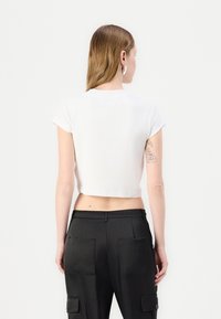White cropped t-shirt with short sleeves and a ribbed texture. The back shows a subtle neckline and a tattoo on the wearer’s arm.