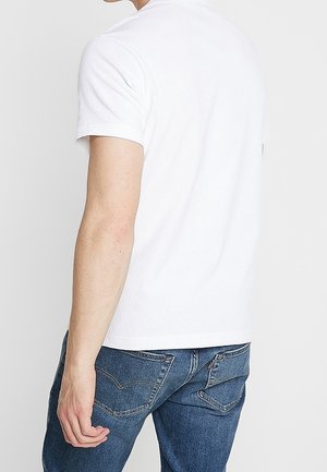 Man wearing white polo shirt and blue jeans, shown from the back with left arm relaxed at his side.