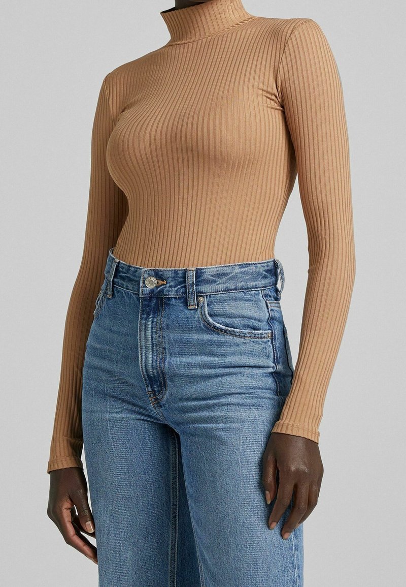 Kvinna klädd i en figurnära ribbad beige polokrage och högmidjade blå denimjeans, stående med armarna avslappnat längs sidorna.