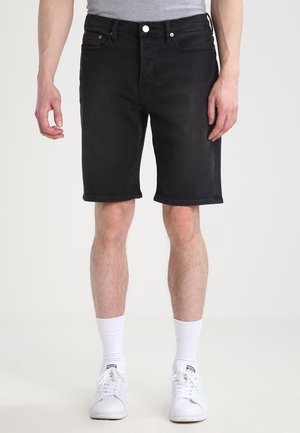 Shorts en denim noir à coupe droite, dotés de cinq poches, d'une fermeture à bouton et d'un ourlet roulé. Associés à des baskets blanches et des chaussettes.