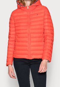 Rød pufferjakke med lynlås foran, tætsiddende hætte og vandret quiltet mønster. Har marineblå detaljer omkring hætten og kraven.