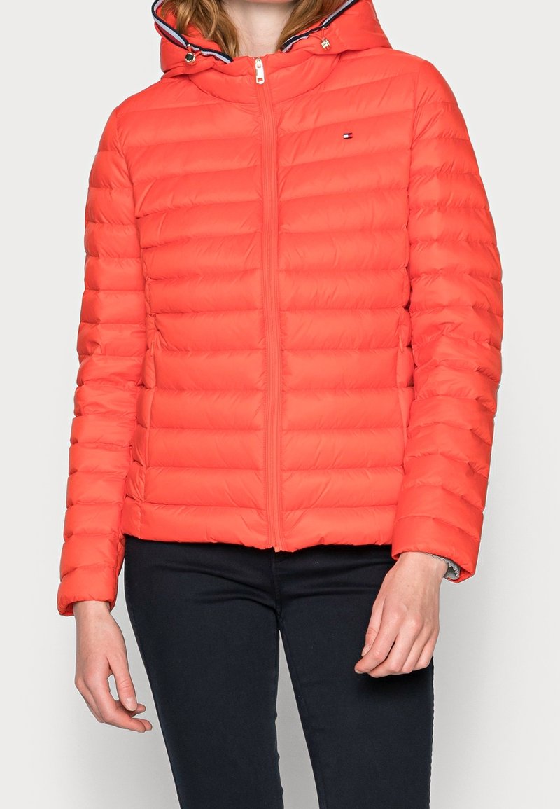 Rød pufferjakke med lynlås foran, tætsiddende hætte og vandret quiltet mønster. Har marineblå detaljer omkring hætten og kraven.