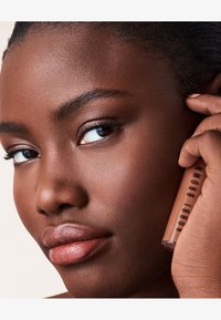 Leppeglansrør holdt av en person med rik brun hud. Glansen har en glatt, glinsende finish og er merket "Bobbi Brown" med fet svart skrift.