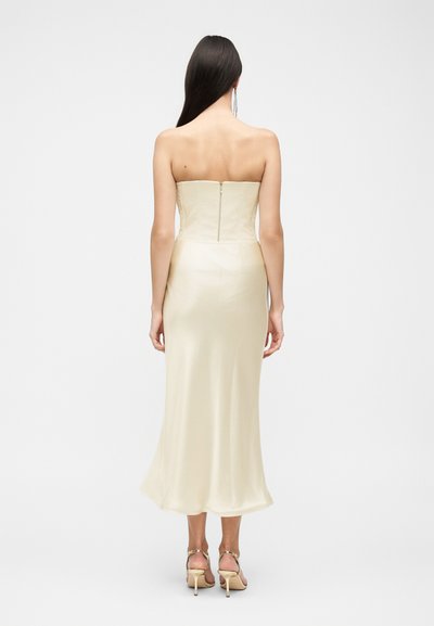 Femme aux longs cheveux foncés portant une robe midi bustier couleur crème et des sandales à talons assorties, debout, de dos.
