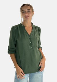 Blusa verde chiusa con bottoni e scollatura a V, tessuto strutturato, maniche con risvolto e taschino sul petto. Vestibilità casual con un design rilassato.
