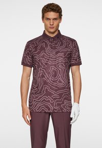 Pikétröja i djup lila med ett topografiskt linjemönster i ljuslila, korta ärmar och en krage med tre knappar.