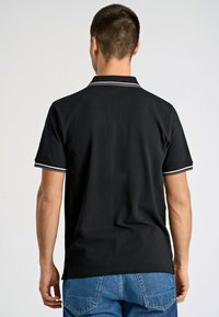 Svart pikétröja med krage, med vita ränder på kragen och ärmarna. Tyget verkar vara slätt och lätt. Denimjeans är synliga.